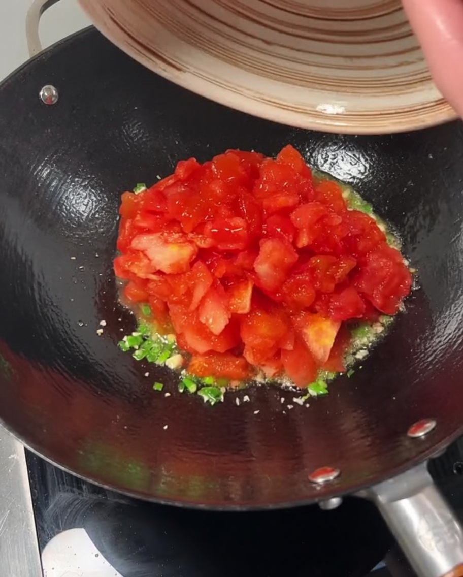 Pomodori a dadini versati da una ciotola in un wok sopra cipollotti e aglio soffritti.