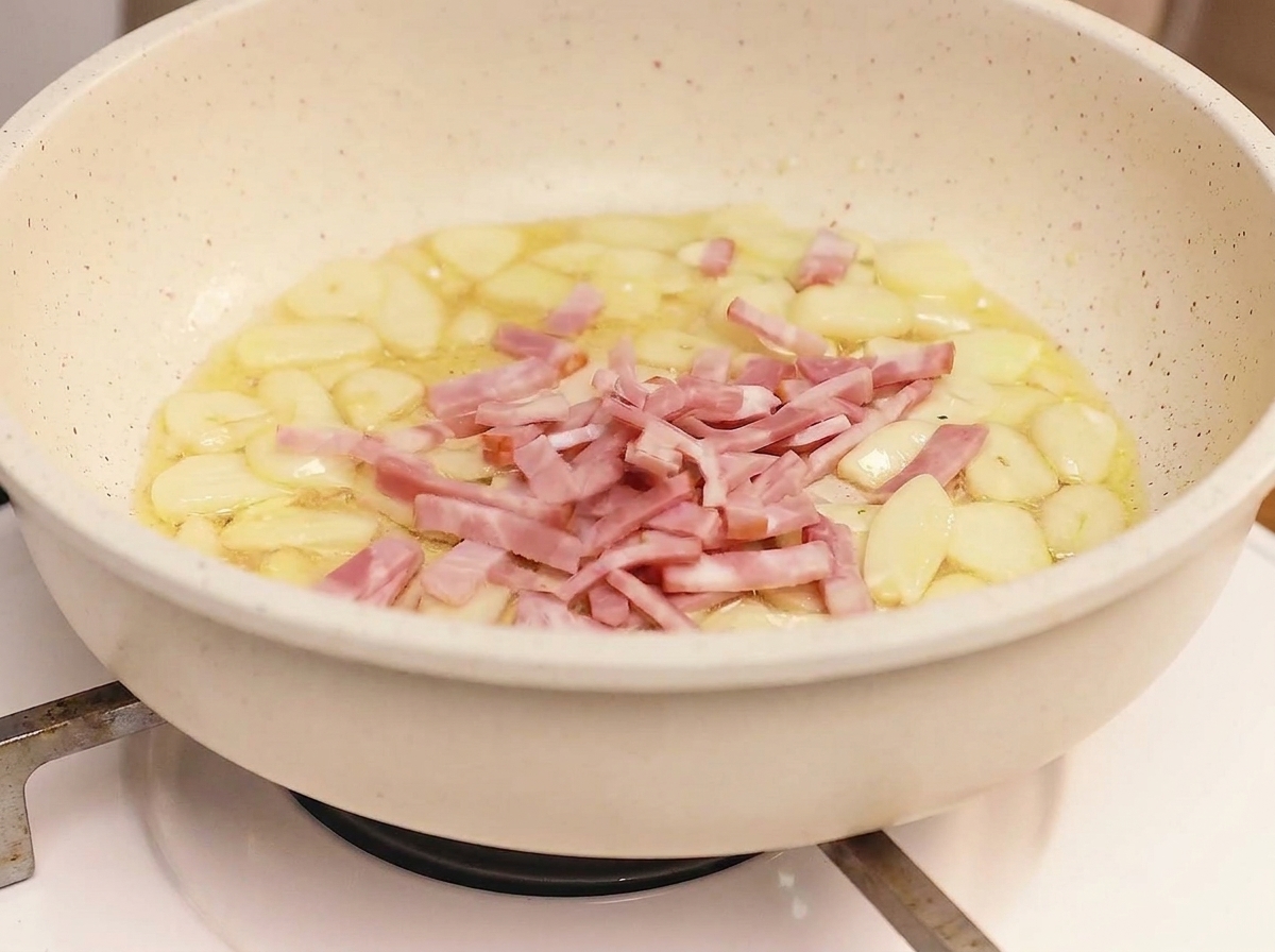 Strisce di pancetta che friggono in padella con aglio, raggiungendo una doratura croccante.