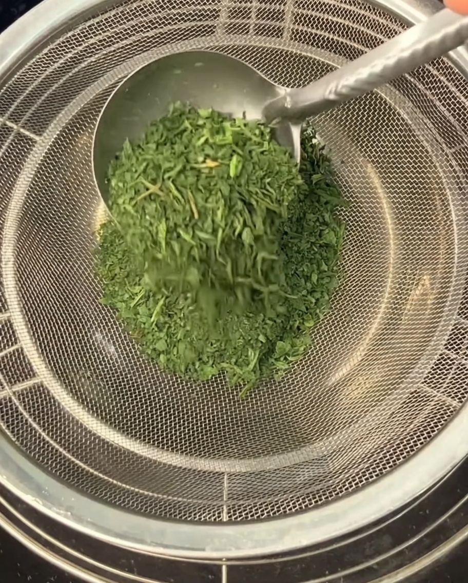 Uso di un cucchiaio di metallo per spingere le foglie di sedano secche e sbriciolate attraverso un setaccio a maglie fini in una ciotola sottostante.