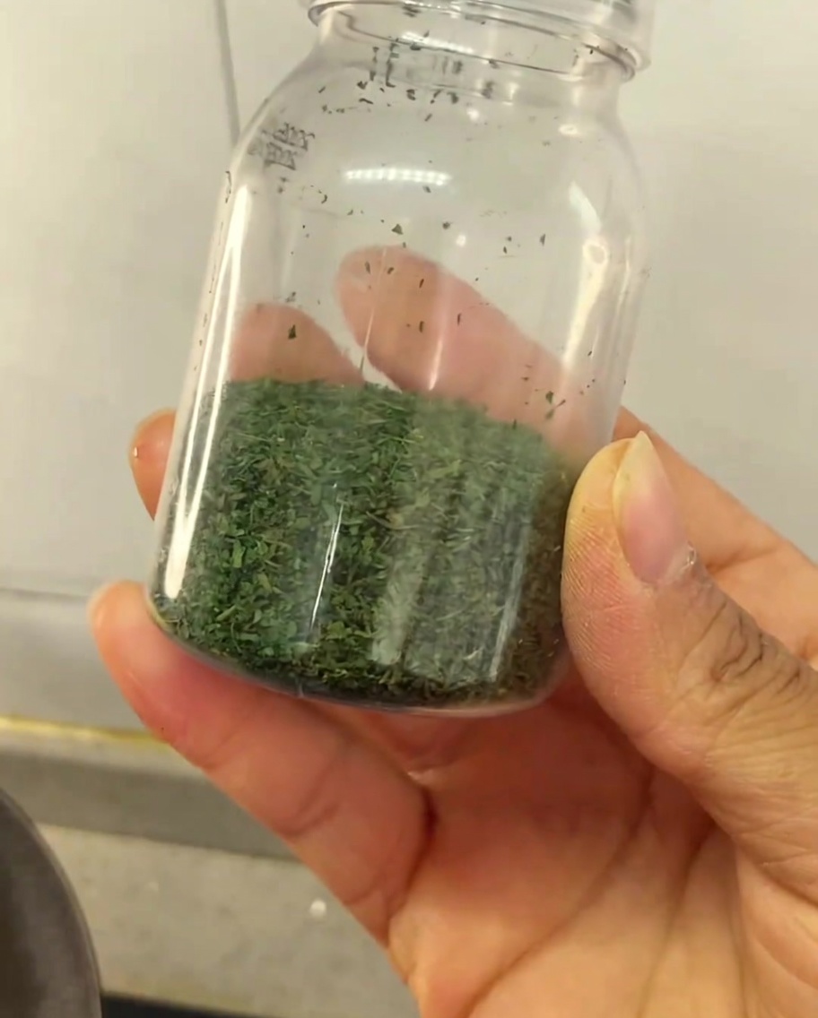 Una mano che tiene un piccolo barattolo di plastica trasparente parzialmente riempito con condimento fatto in casa a base di foglie di sedano essiccate verdi.