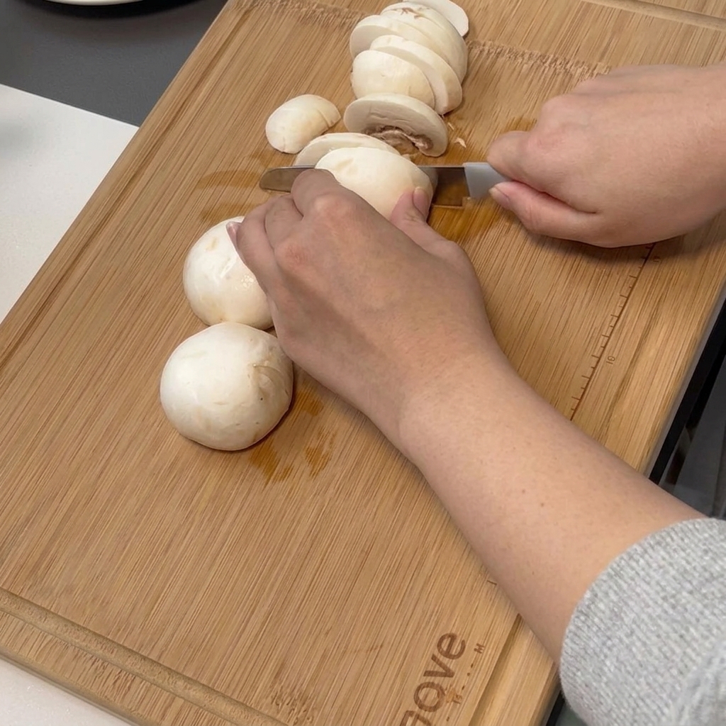 Mano che affetta funghi champignon su un tagliere di bambù.