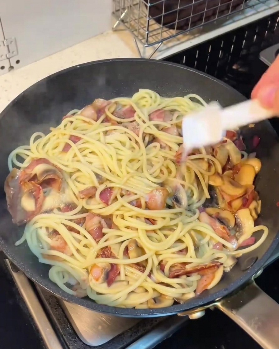 Una mano che aggiunge pepe nero macinato sopra gli spaghetti in padella.