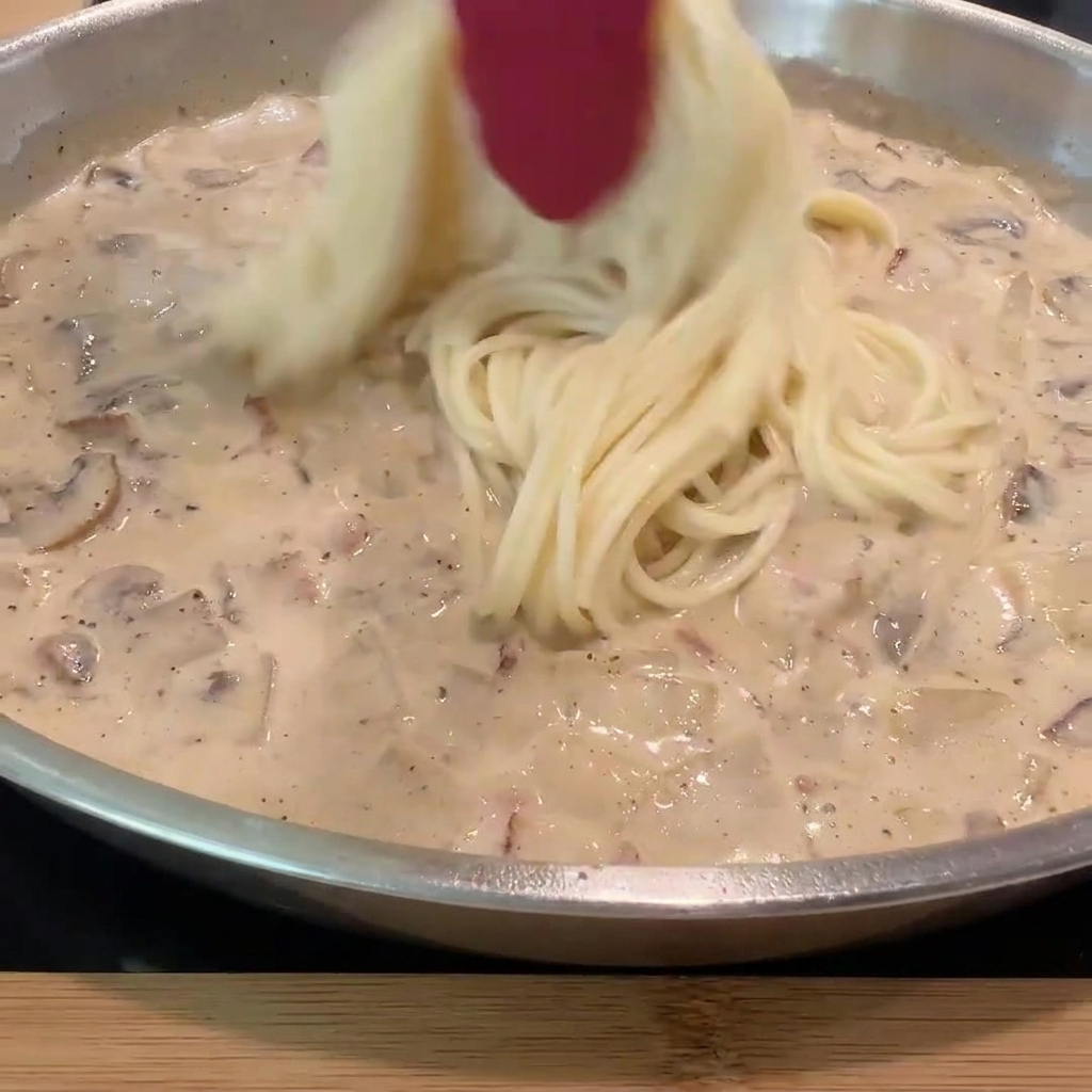 Spaghetti precotti aggiunti alla padella con salsa cremosa e ingredienti.