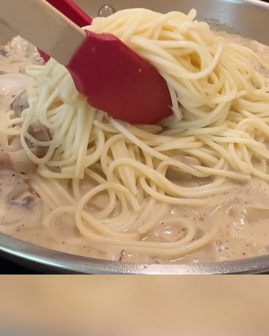 Spaghetti saltati con salsa cremosa, pancetta e funghi in una padella usando pinze rosse.