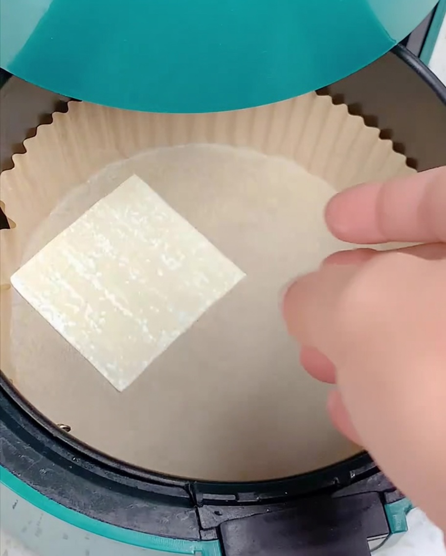 Una mano posiziona un foglio di pasta wonton quadrato nel cestello della friggitrice ad aria foderato con carta forno.