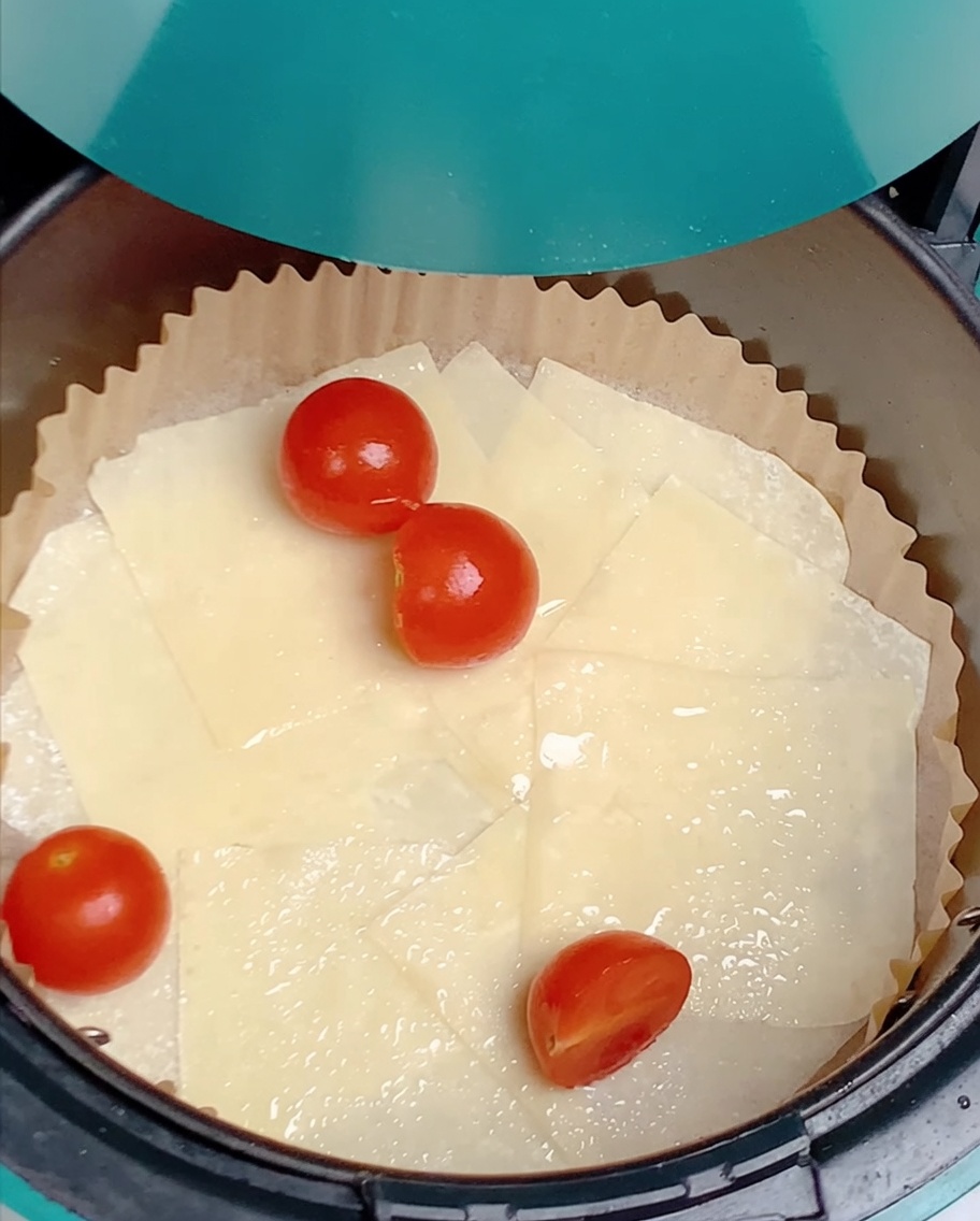 Pomodorini tagliati a metà sparsi sopra i fogli di wonton oleati nel cestello della friggitrice ad aria.