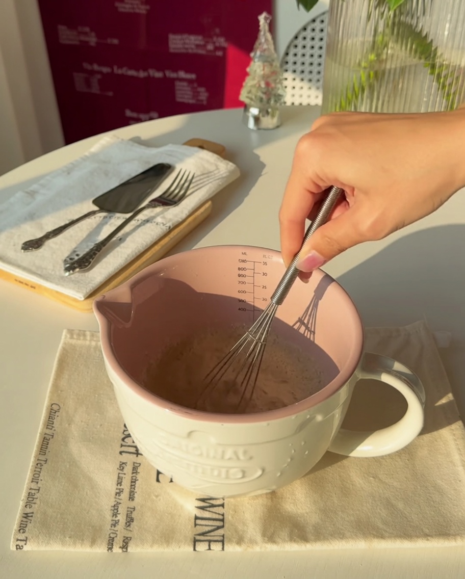 Primo piano di una mano che mescola il lievito nellacqua tiepida dentro un misurino rosa.