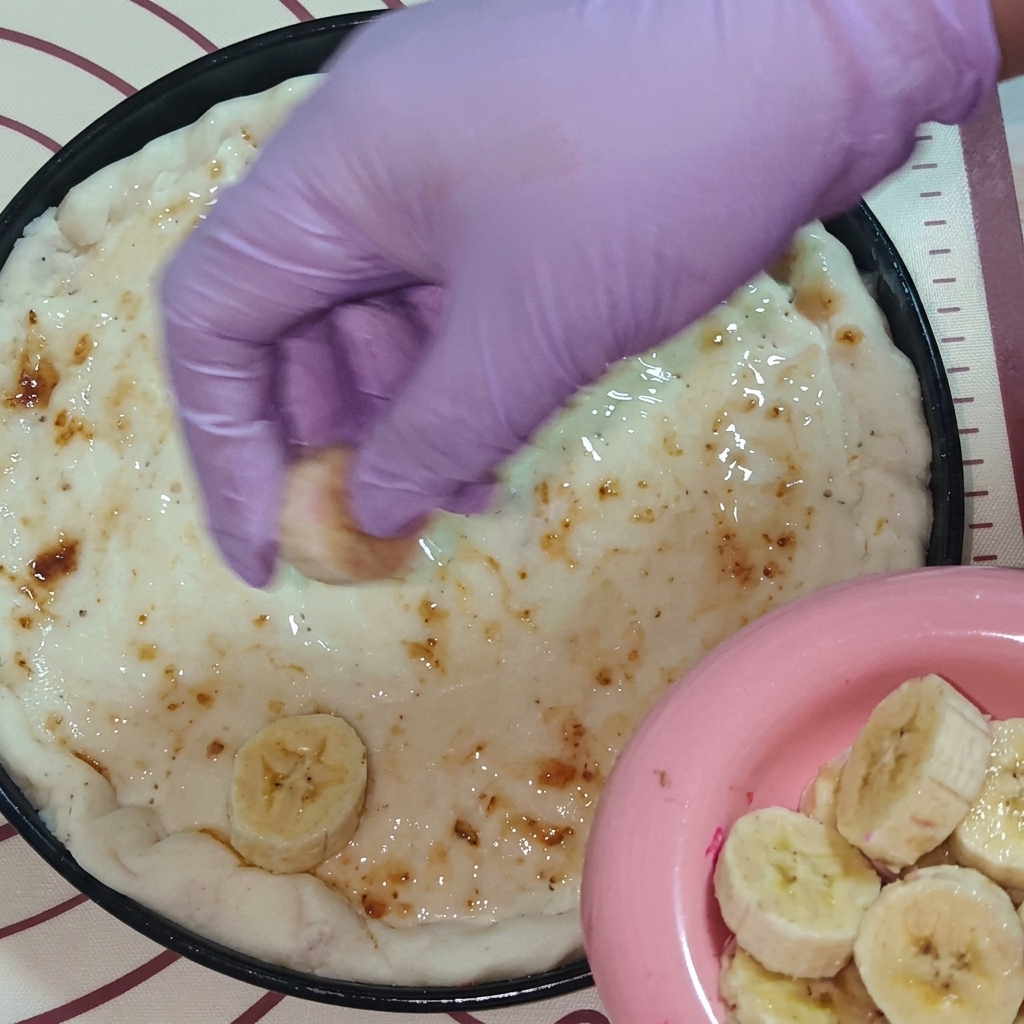 Una mano con guanto viola che posiziona fette di frutta fresca, inclusi banana e pitaya, sulla pizza.