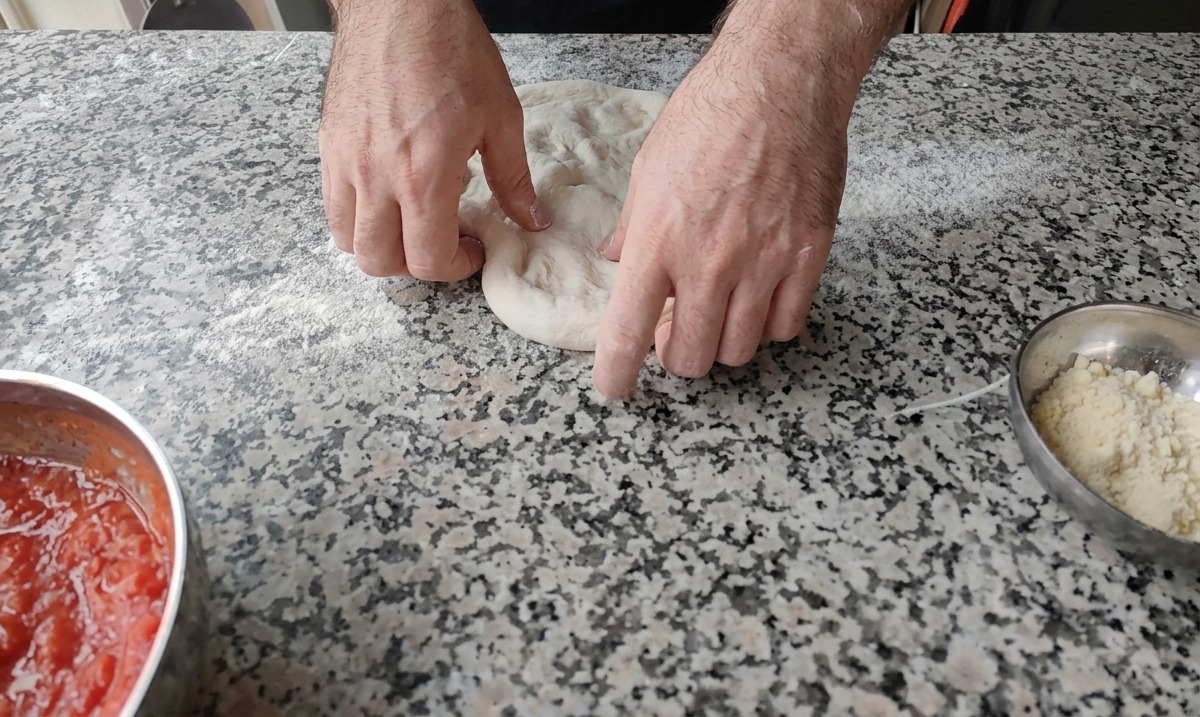 Due mani che premono al centro di un panetto di impasto per pizza su un piano di marmo leggermente infarinato.