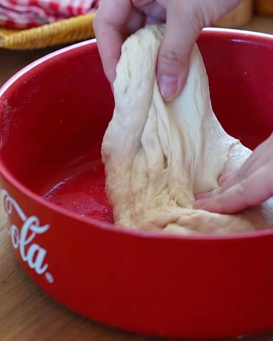 Primo piano di mani che eseguono la tecnica stretch-and-fold su un impasto soffice in una ciotola rossa.