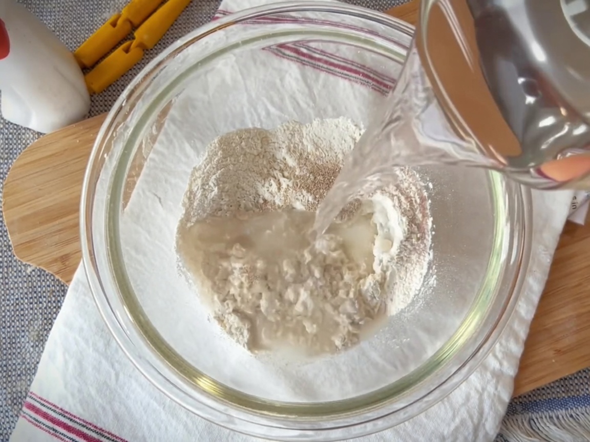 Una ciotola di vetro contenente un mix di farina, lievito, sale e acqua, mescolati con una spatola.