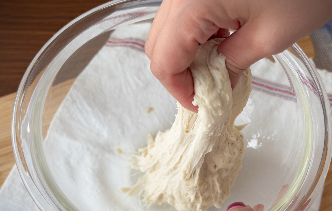 Due mani bagnate che allungano e ripiegano limpasto ad alta idratazione allinterno di una ciotola.