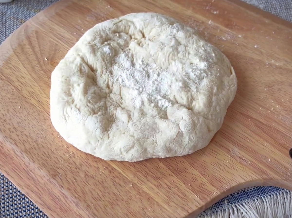 Un panetto di pasta per pizza steso su una superficie in legno, con un cornicione spesso ben definito.