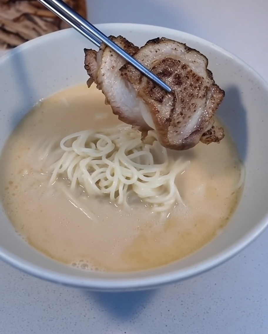 Una fetta di Char Siu scottato posizionata su una ciotola di ramen con delle pinzette.