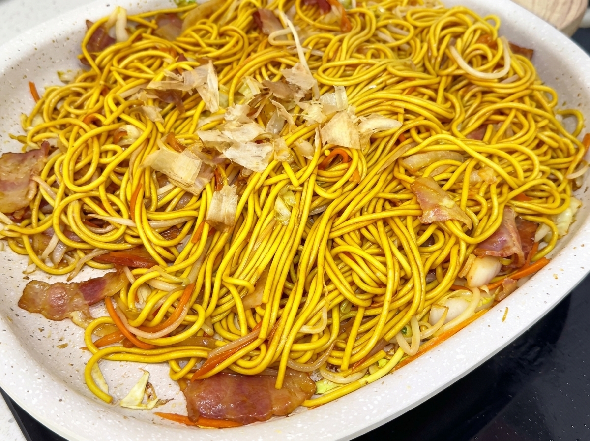 Una padella piena di spaghetti saltati coperti da una pioggia di fiocchi di bonito.