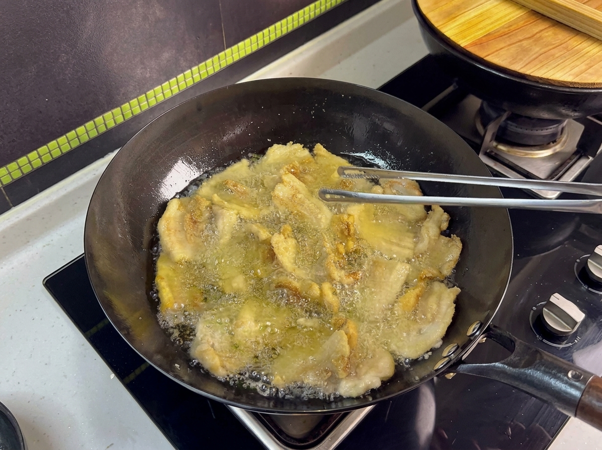 Diverse fette di pancetta che friggono in un wok, girate con pinze di metallo.
