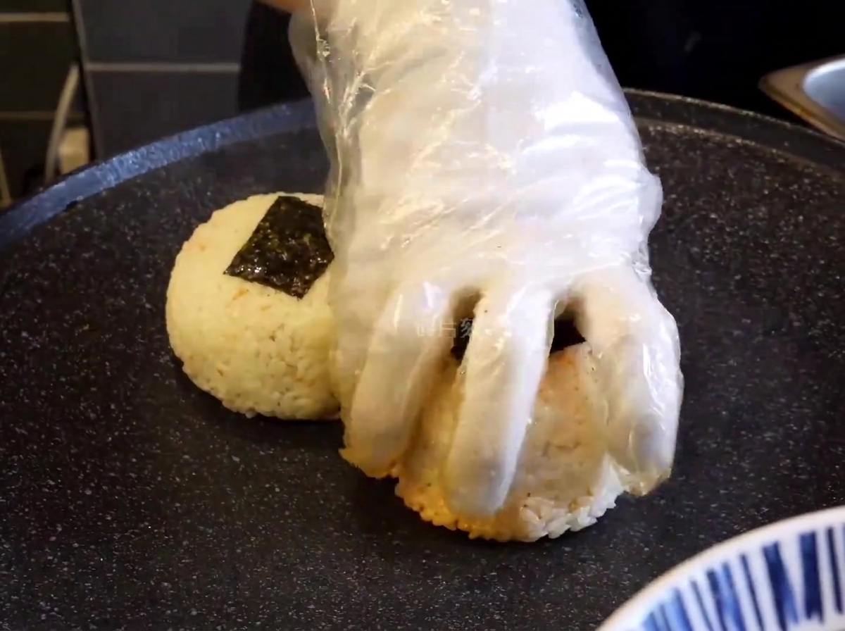 Mano guantata che preme le polpette di riso su una piastra nera per farle rosolare.