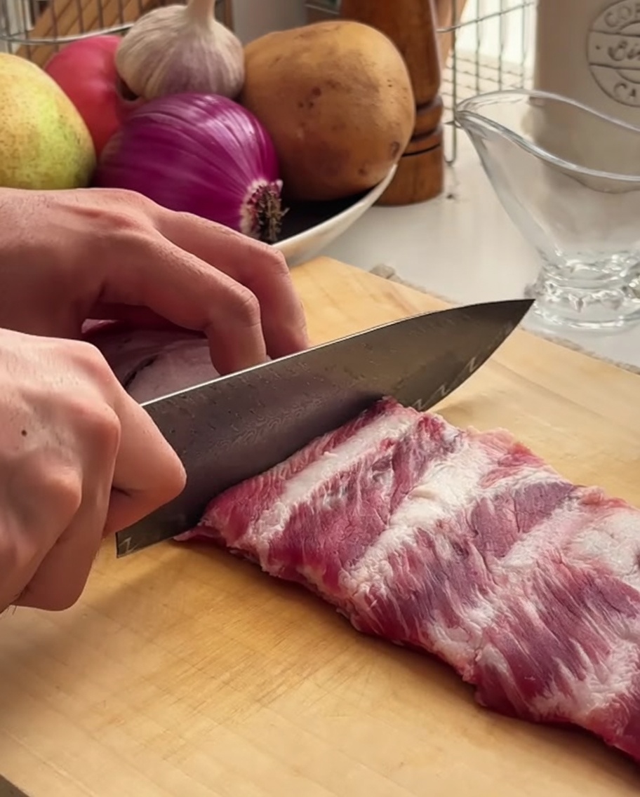 Un primo piano di uno chef che usa un coltello affilato per porzionare un rack di costine di maiale su un tagliere di legno.