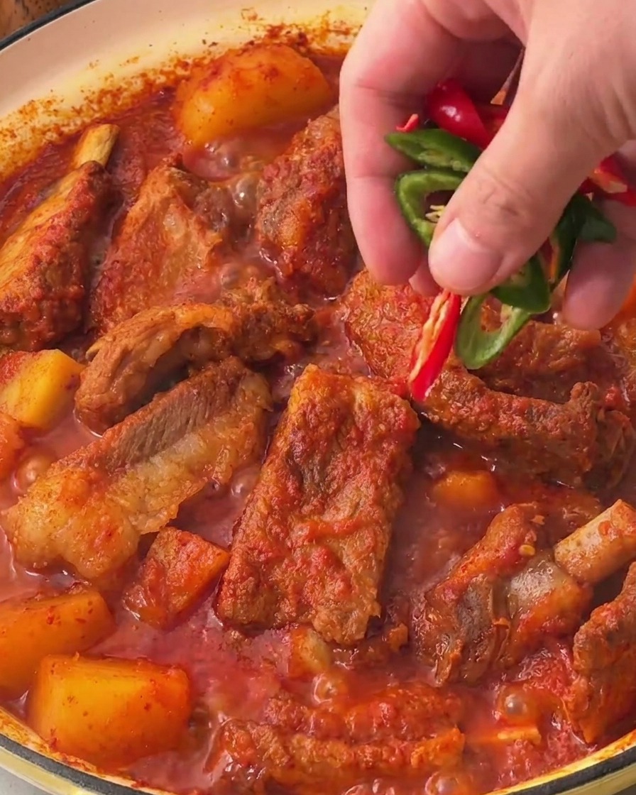 Una mano che sparge fette di peperoncino fresco, rosso e verde brillante, sopra la superficie delle costine di maiale brasate piccanti coreane finite.