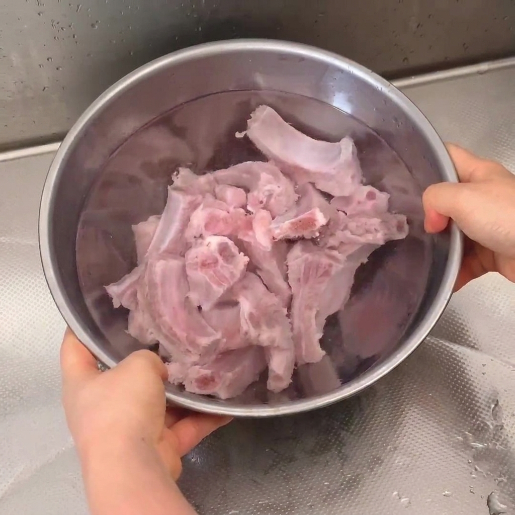Costine di maiale fresche in ammollo e sciacquate in una ciotola di acciaio inox sotto il rubinetto della cucina.