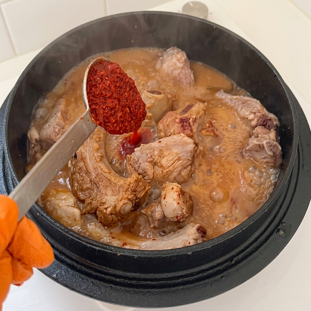 Cucchiaio che aggiunge peperoncino in polvere e aglio tritato in una pentola contenente costine di maiale e condimento liquido.