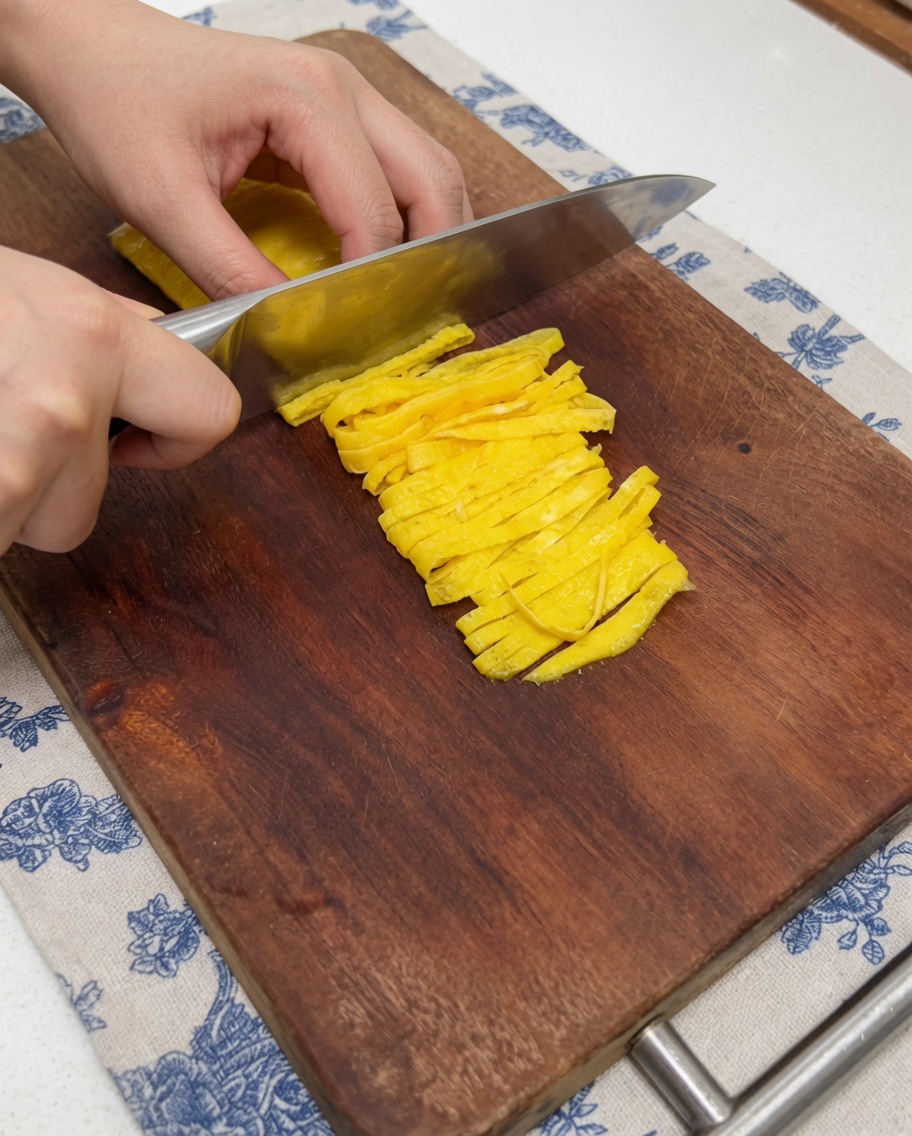 Primo piano di una mano che usa un coltello da cucina per tagliare una frittatina di uova cotta e gialla in strisce sottili su un tagliere di legno.