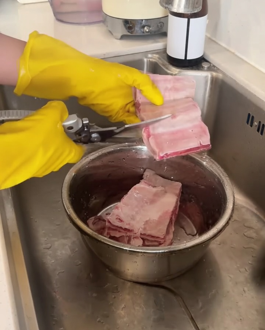 Un paio di mani con guanti gialli usano le forbici da cucina per tagliare costine di manzo crude su una ciotola di metallo.