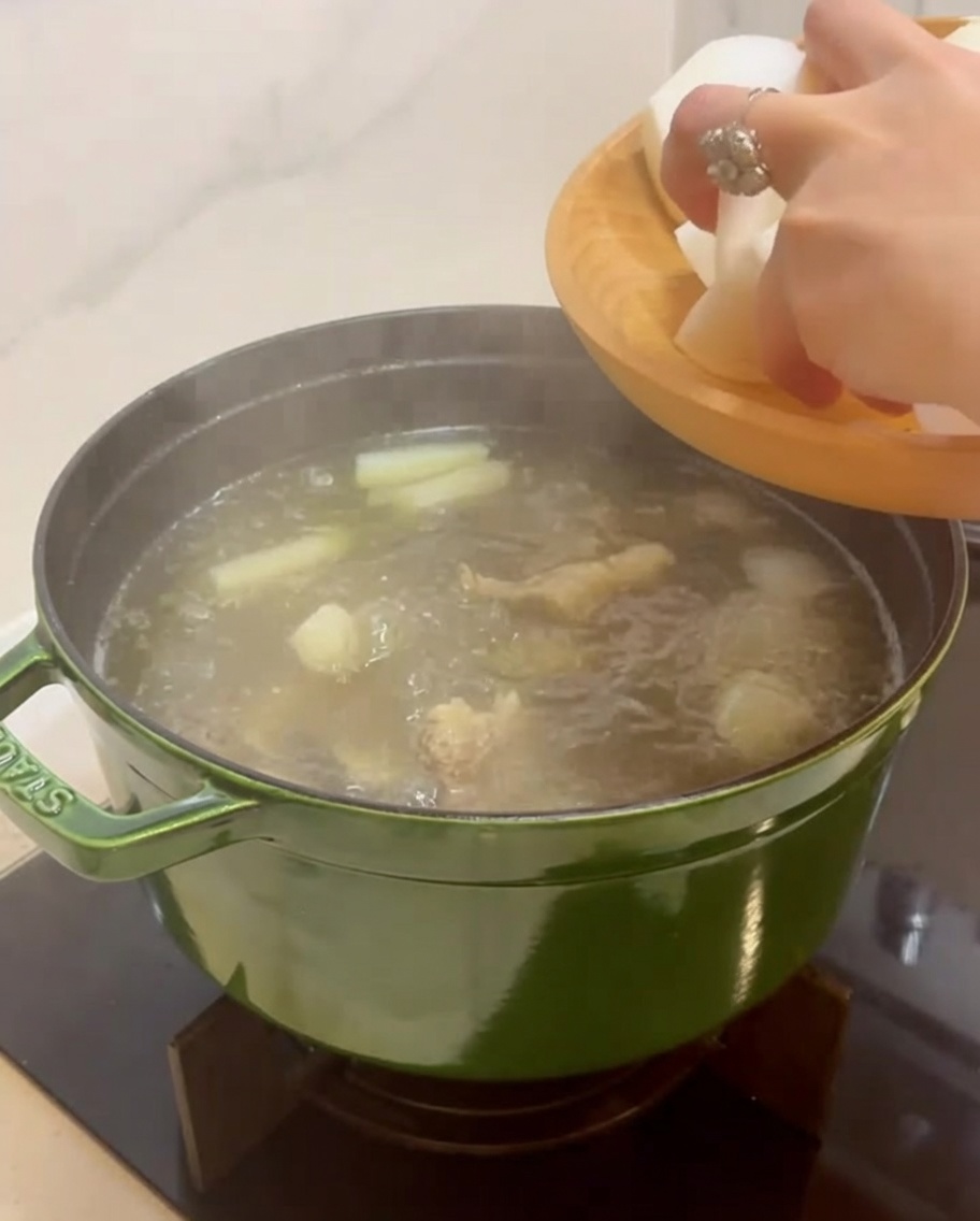 Mano che aggiunge grandi pezzi bianchi di ravanello coreano sbucciato in una pentola di zuppa di manzo che sobbolle.