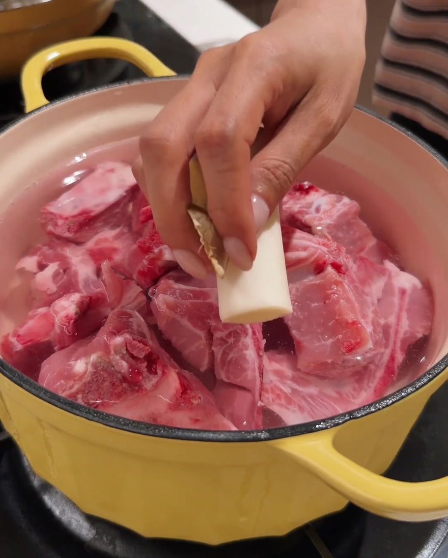 Una mano che posiziona un pezzo di zenzero in una pentola contenente spina di maiale e acqua.