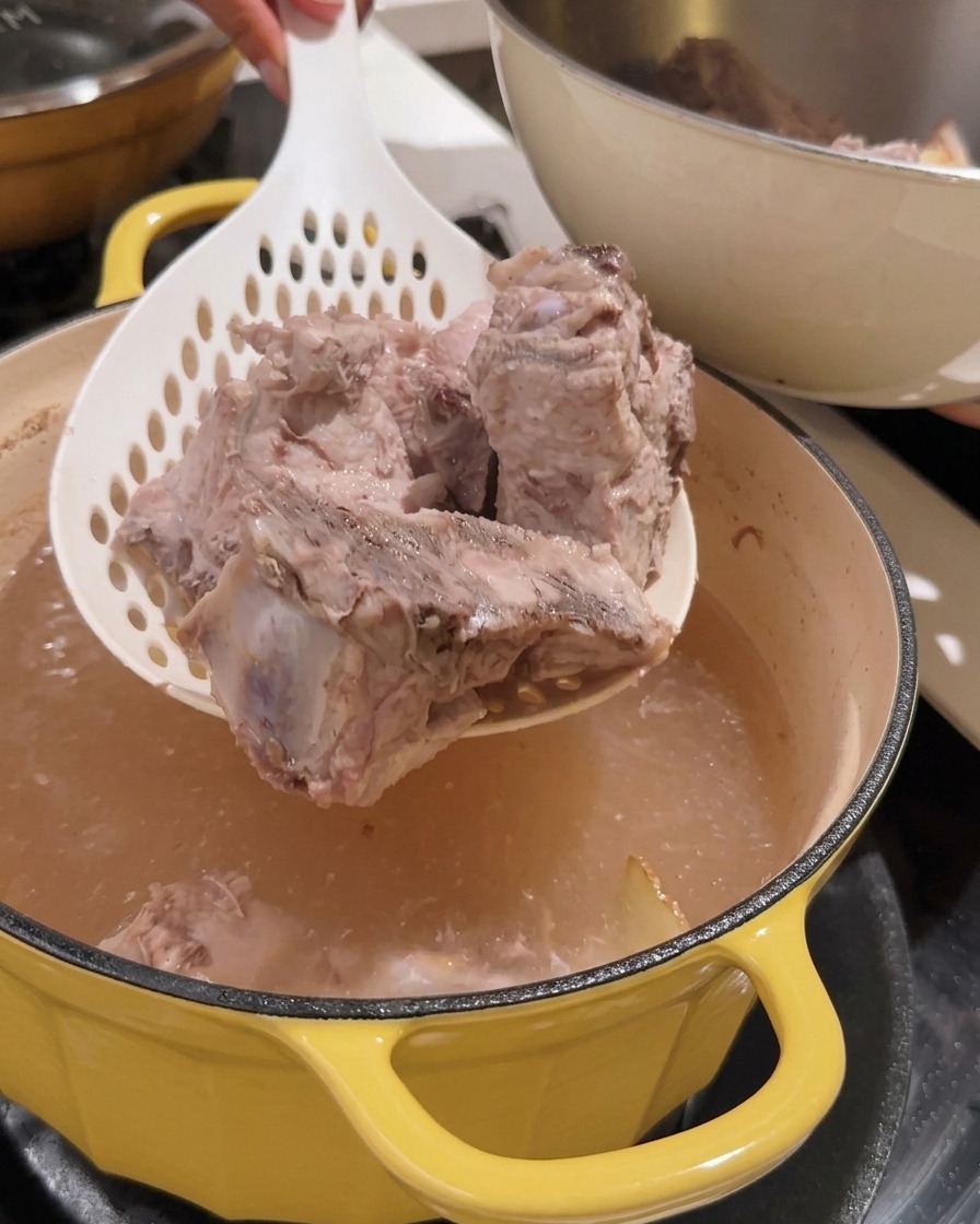 Scolapasta che solleva pezzi di spina di maiale sbollentati da una pentola.