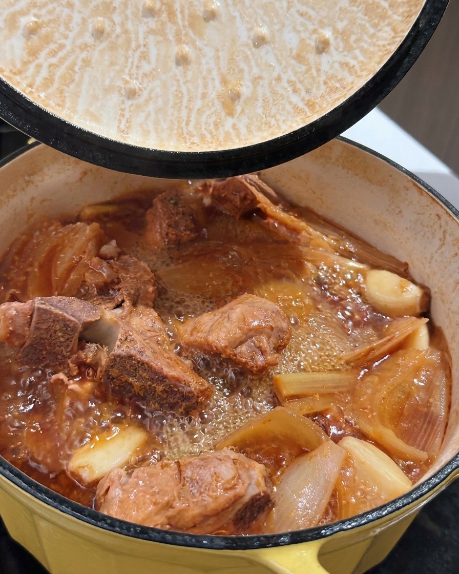 Una pentola di costine di maiale brasate e ribollenti con brodo ricco mentre viene sollevato il coperchio.