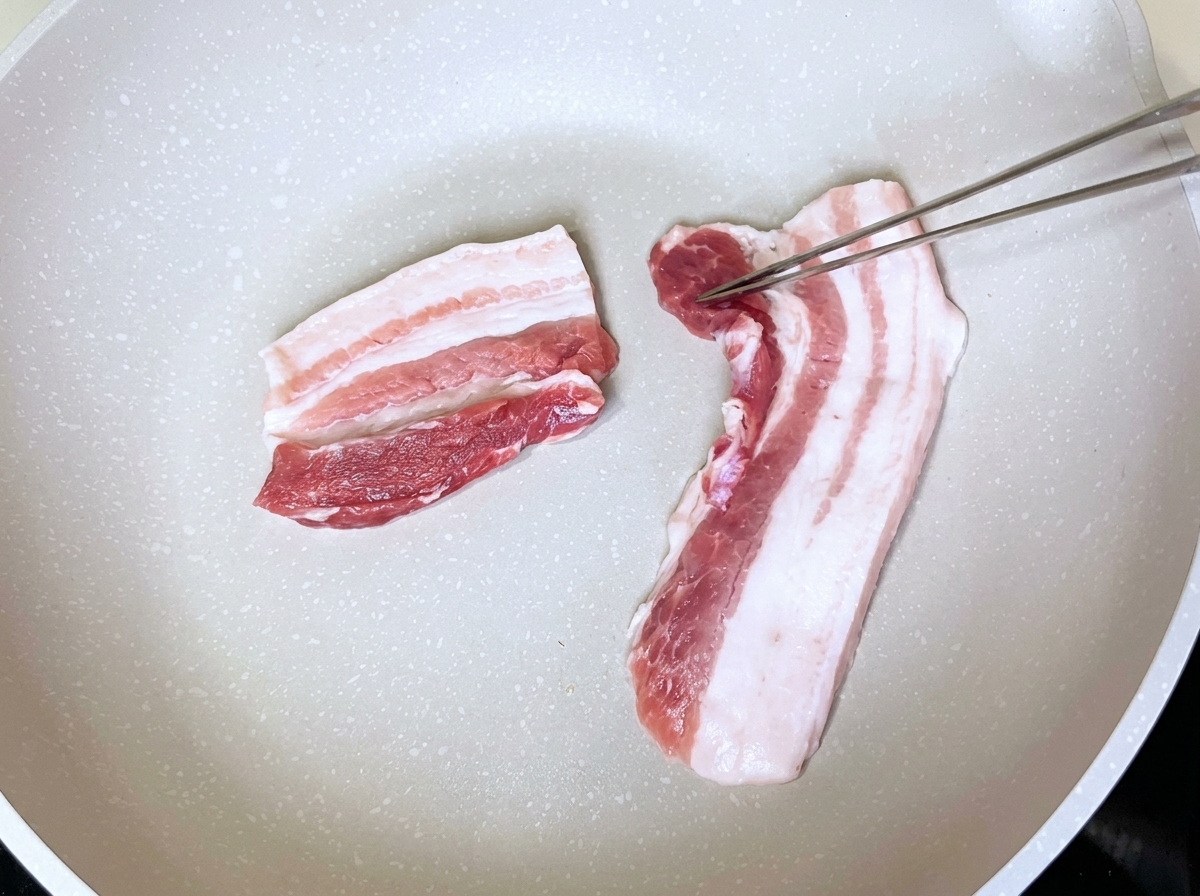 Fette di pancetta in una padella bianca asciutta gestite con pinze metalliche.