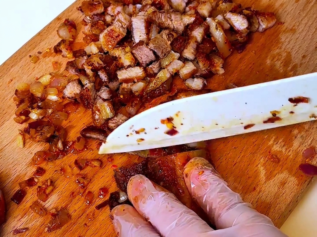 Un coltello trita il maiale cotto e le cipolle su un tagliere in legno, con mani che indossano guanti protettivi.