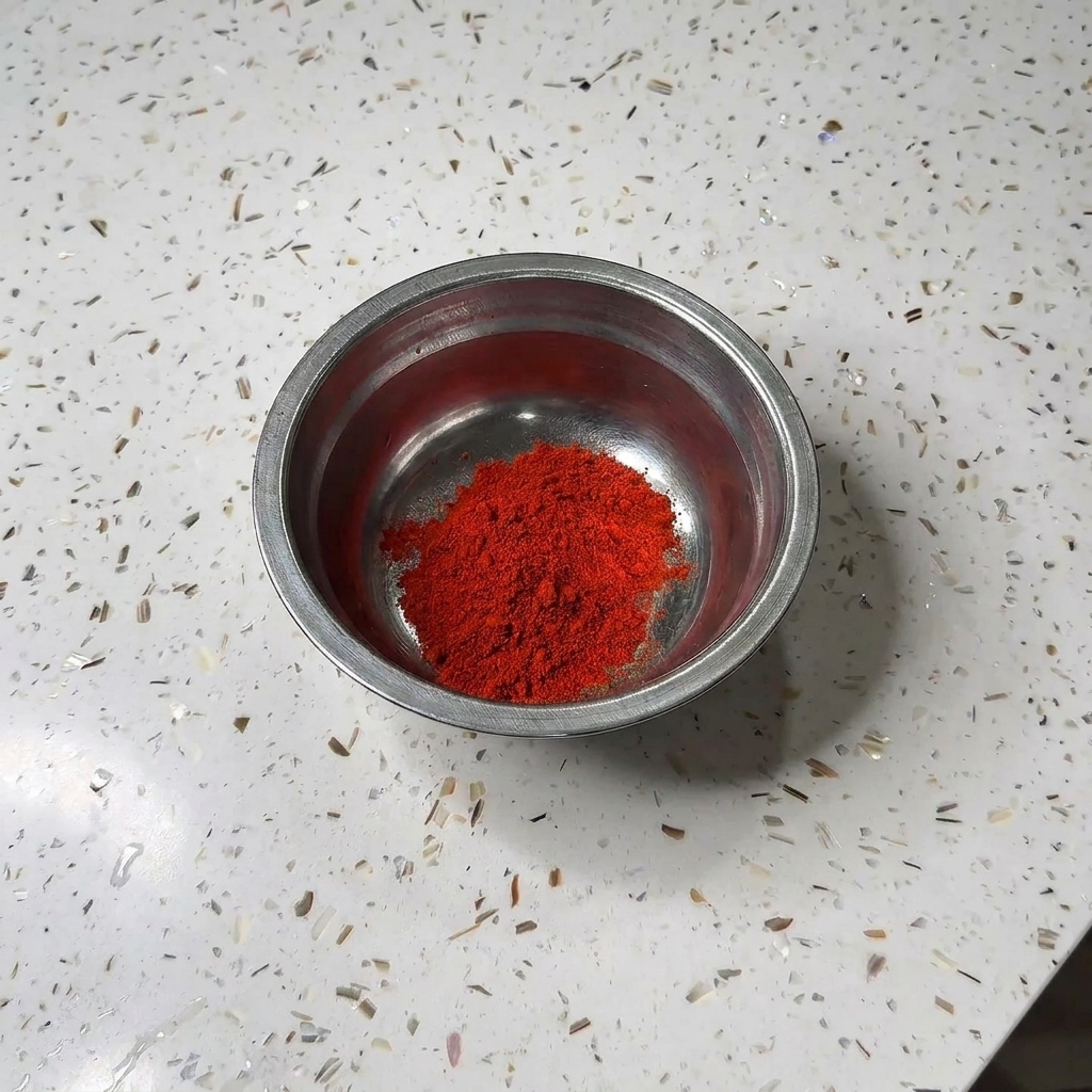Una piccola ciotola di metallo contenente un mucchietto di peperoncino in polvere rosso e zucchero bianco su un piano di lavoro in granito.