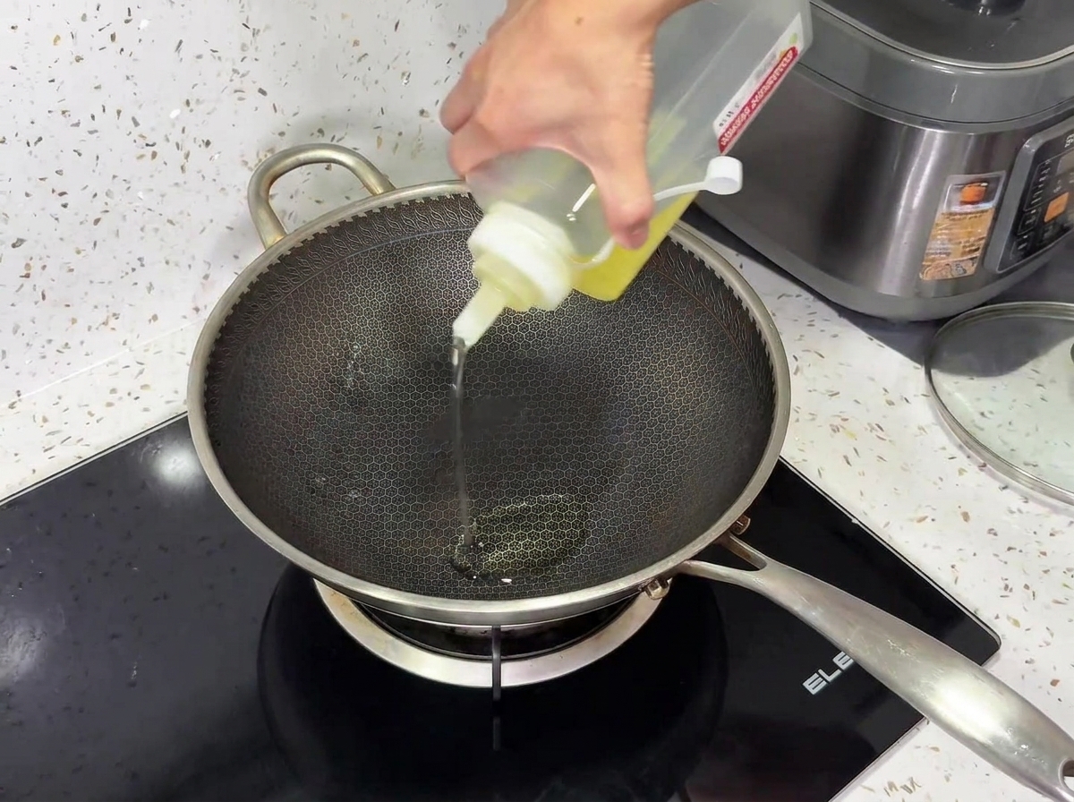 Olio da cucina versato da una bottiglia dosatrice in un wok di metallo texturizzato su un piano cottura a induzione nero.