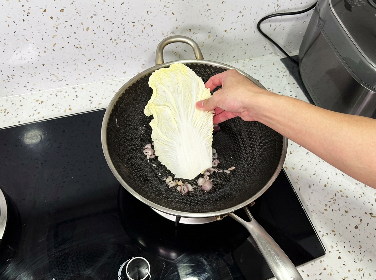 Una mano che posiziona una grande foglia di cavolo napa in un wok contenente cipolle cotte.