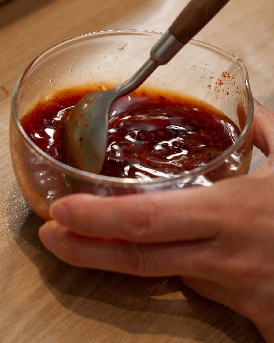 Un cucchiaio che mescola una miscela di salsa per hot pot marrone rossastra in una ciotola di vetro.