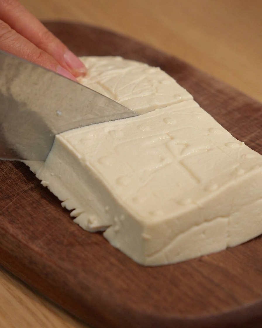 Un coltello affilato che taglia il tofu sodo in blocchi rettangolari ordinati su un tagliere di legno.