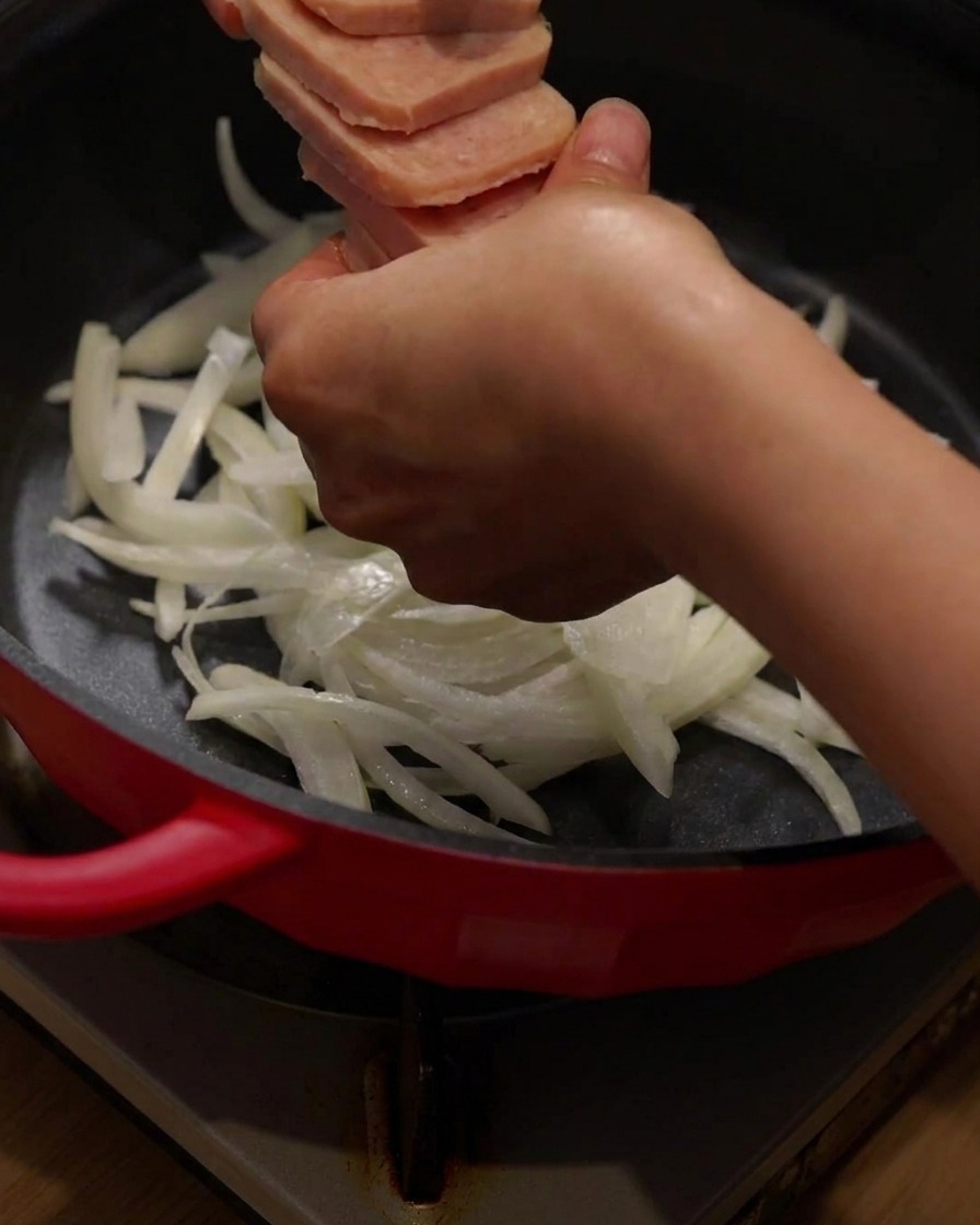 Disposizione di carne in scatola affettata e pancetta sopra le cipolle nella pentola di cottura rossa.