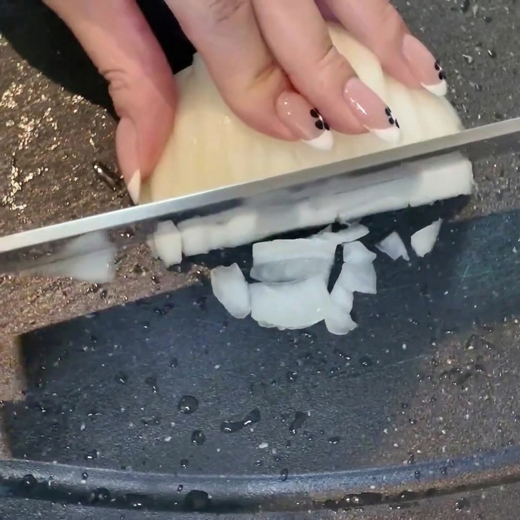 Una mano che taglia una cipolla bianca a cubetti su un tagliere scuro, con aglio tritato accanto.
