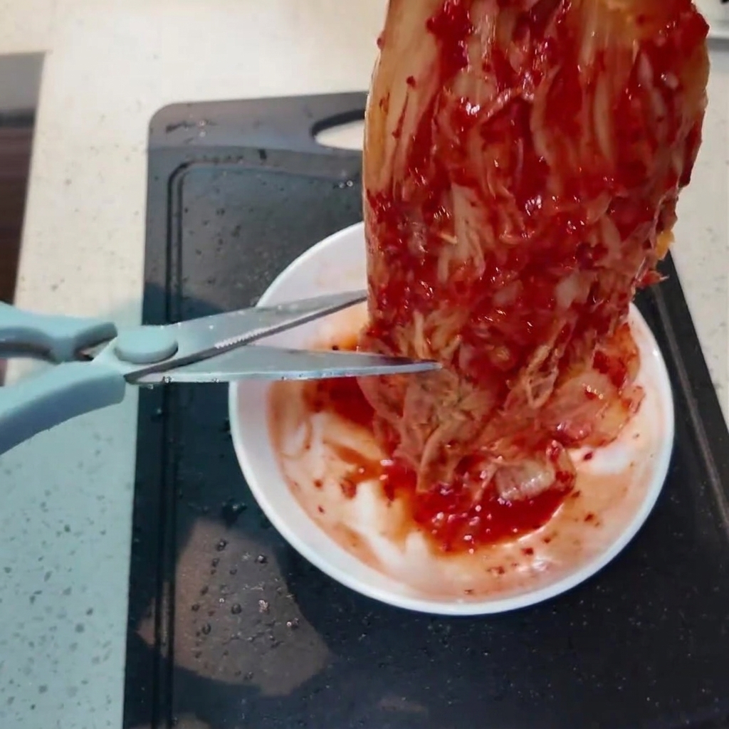 Un pezzo di vivace kimchi rosso tagliato in pezzi più piccoli con delle forbici da cucina sopra un piattino bianco.