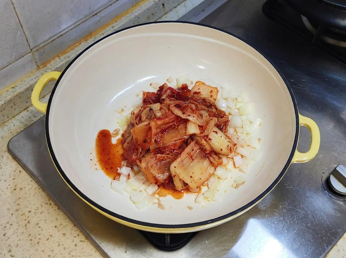 Una porzione di kimchi rosso fermentato sopra delle cipolle soffritte in una padella bianca.