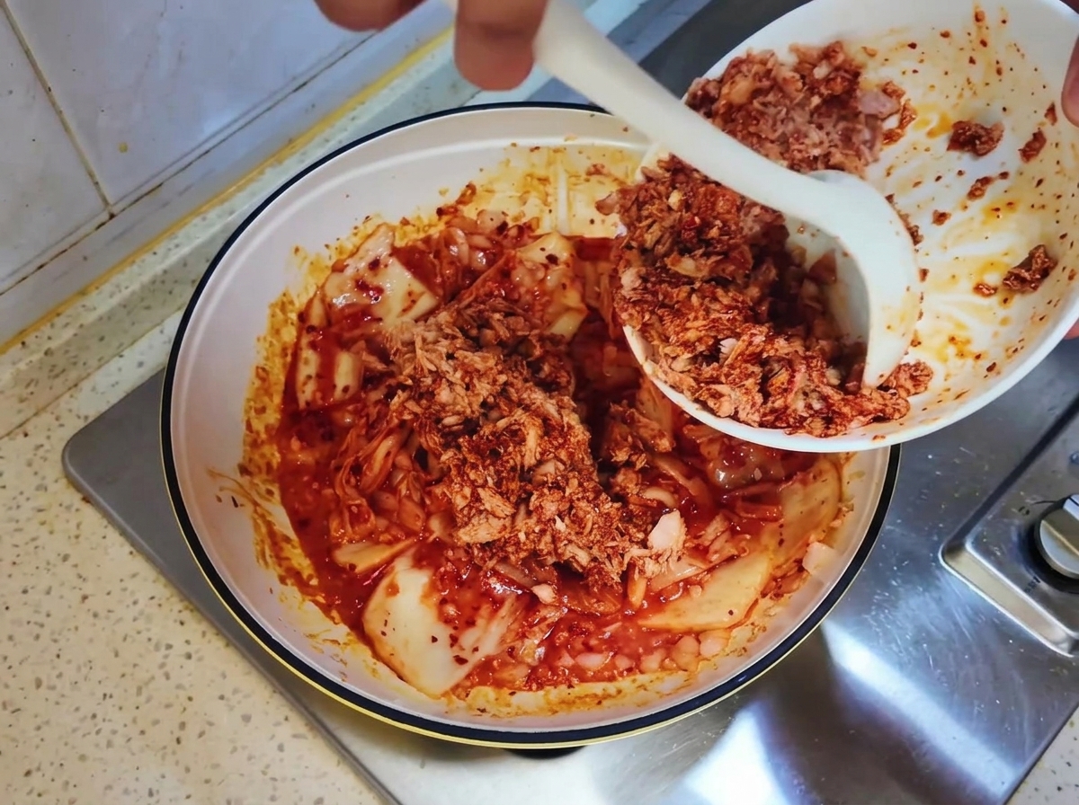 Il composto di tonno in scatola viene raschiato da una ciotola bianca nella padella con il kimchi usando un cucchiaio bianco.