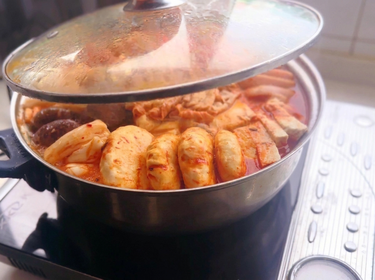 Un coperchio di vetro viene sollevato da una pentola calda di metallo piena di ingredienti disposti come carne affettata, tofu, funghi, torte di riso e gnocchi in un brodo rosso piccante su una piastra a induzione.