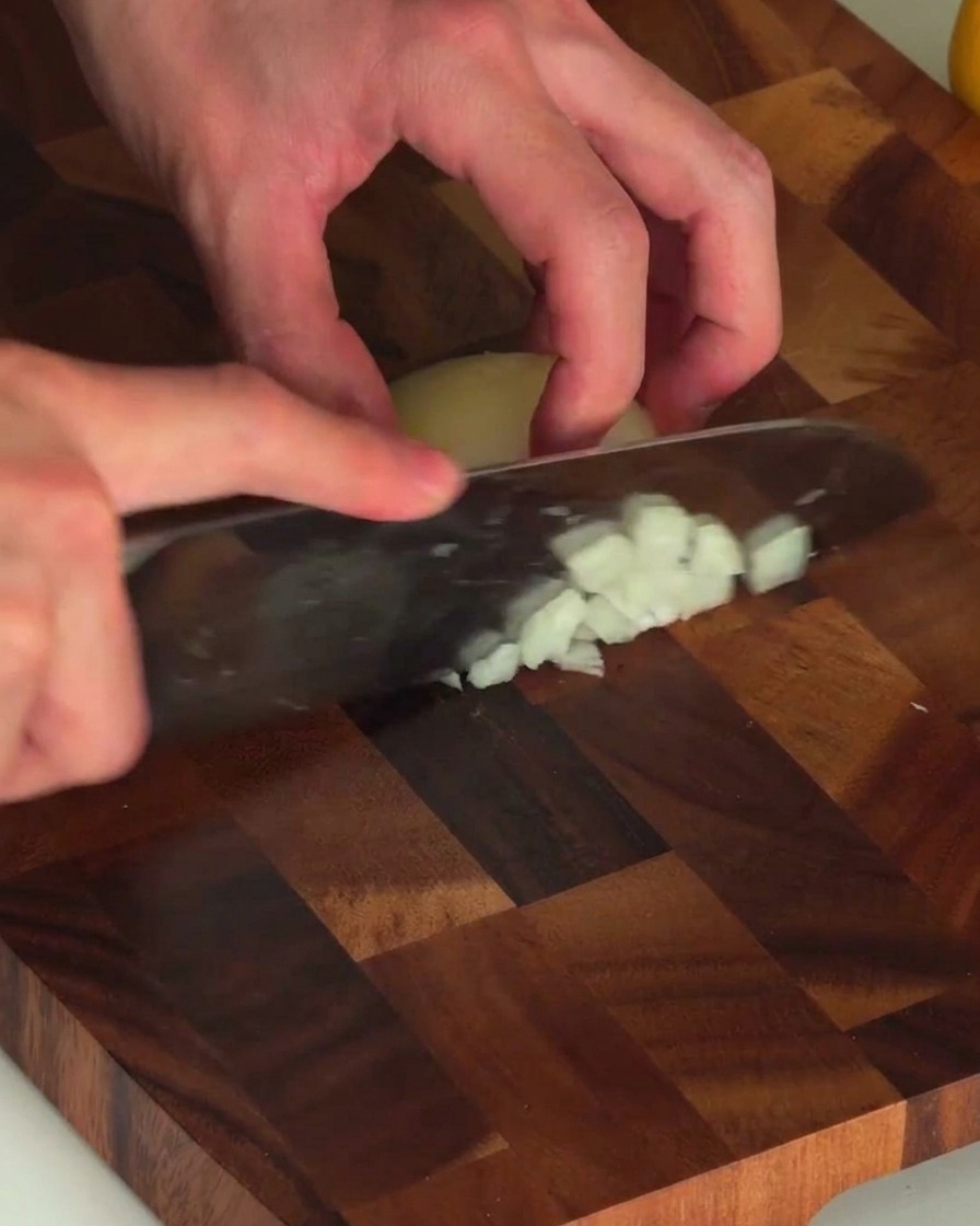 Mani che tengono un coltello e tritano finemente una cipolla bianca su un tagliere di legno.