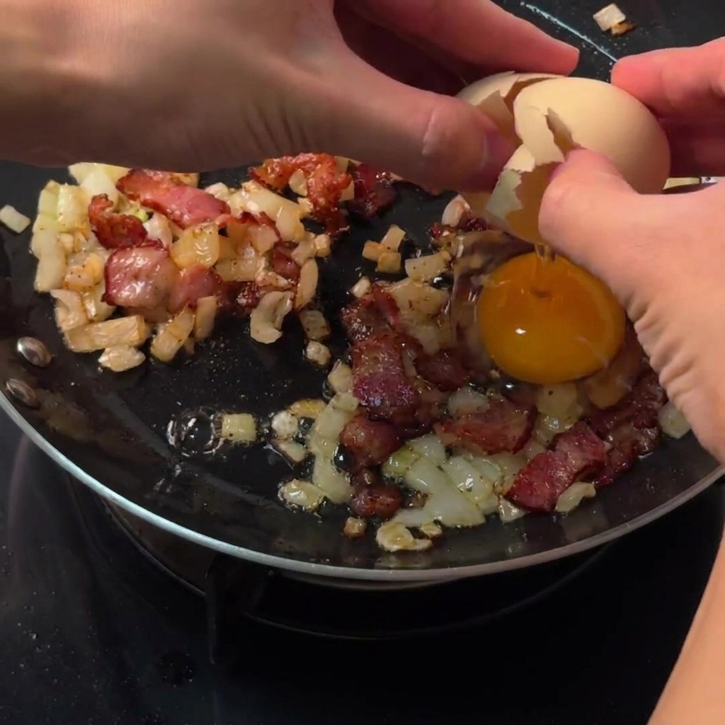 Mani che rompono un uovo crudo direttamente nello spazio circolare libero tra pancetta e cipolle in padella.