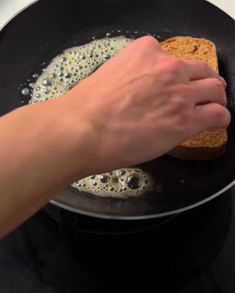 Una mano che posiziona una fetta di pane integrale sul burro fuso e sfrigolante in una padella calda.