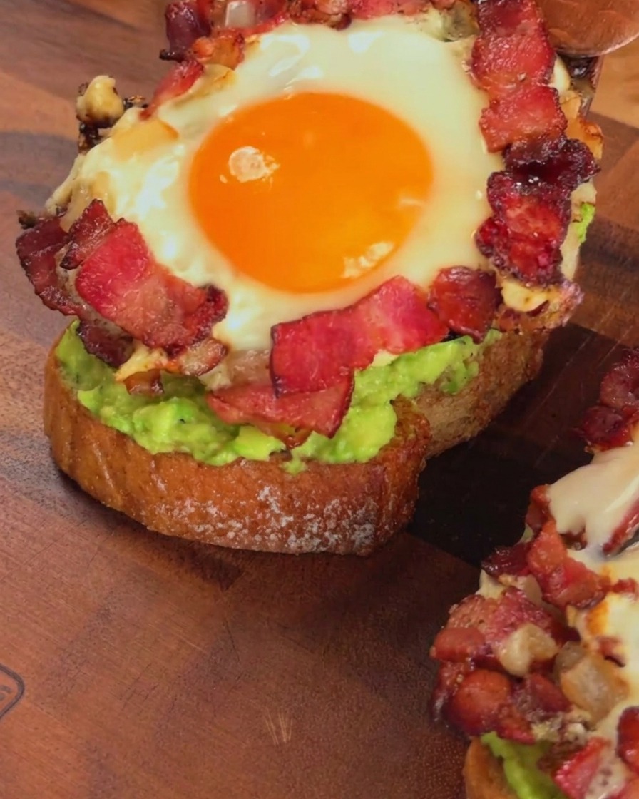 Un anello di uovo e pancetta cotto viene posizionato sullavocado toast con una spatola di legno.