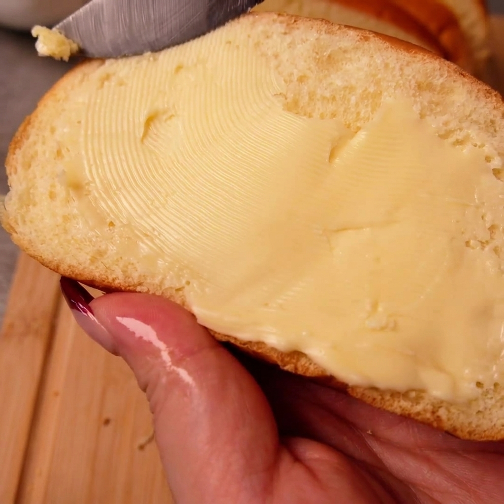 Una mano che tiene una fetta di pane bianco mentre spalma uniformemente il burro sulla superficie con un coltello.