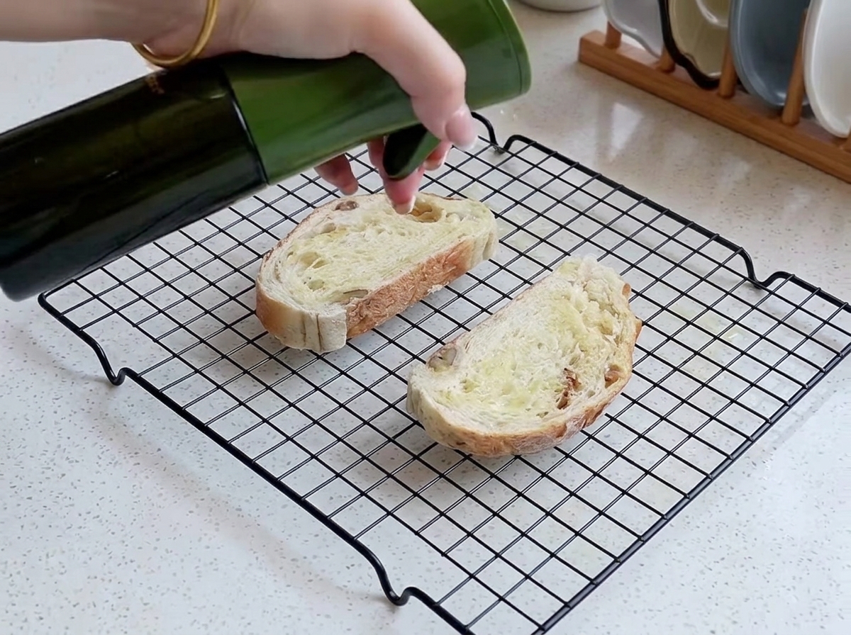 Una mano che spruzza un sottile strato di olio su due fette di pane rustico disposte su una griglia di raffreddamento.